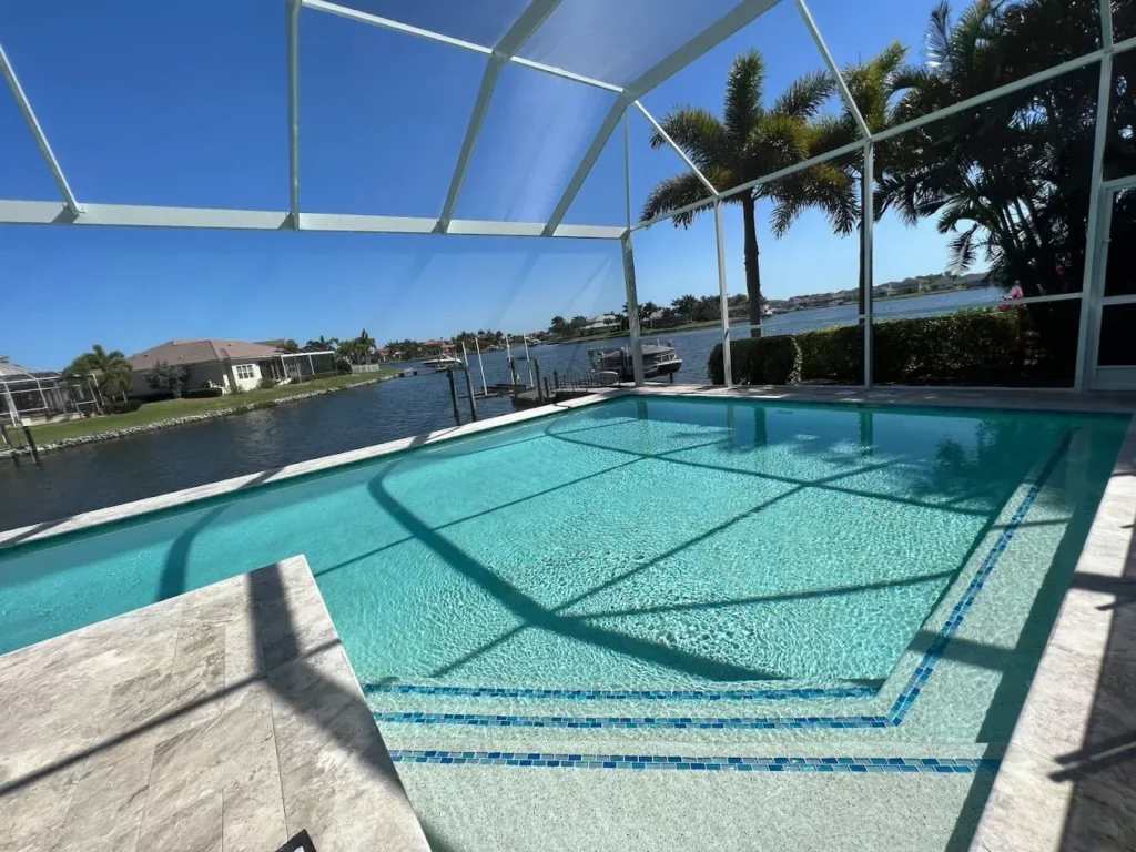 A screened, multilevel geometric pool with mosaic tile steps overlooking a canal with boats docked nearby.