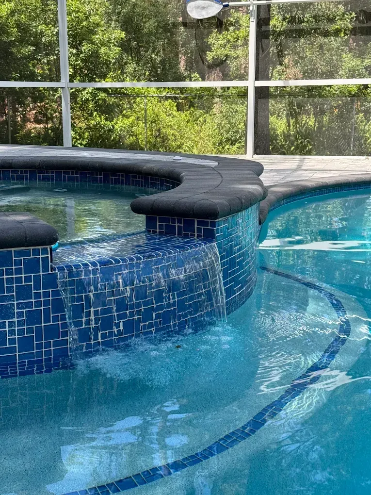 A tiled raised spa spills water into a pool under a screened enclosure.