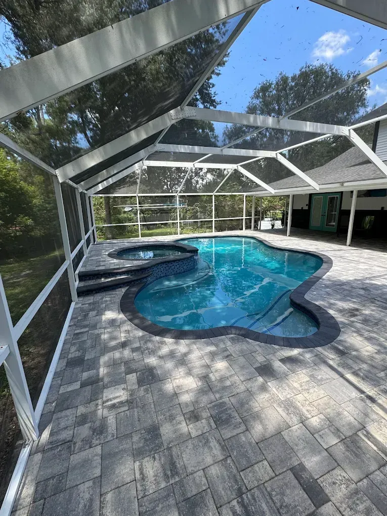 A freeform pool with an attached spa under a screened patio enclosure with paver decking.