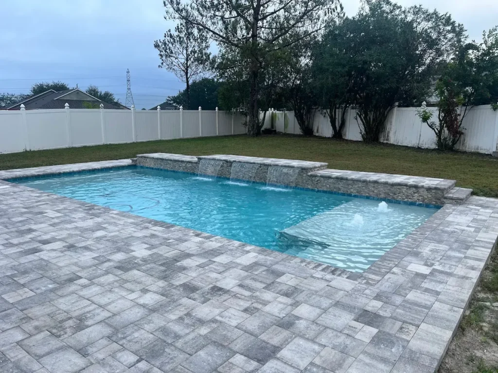 A rectangular backyard pool with a raised stone wall featuring four water sheer spillways and paver decking.