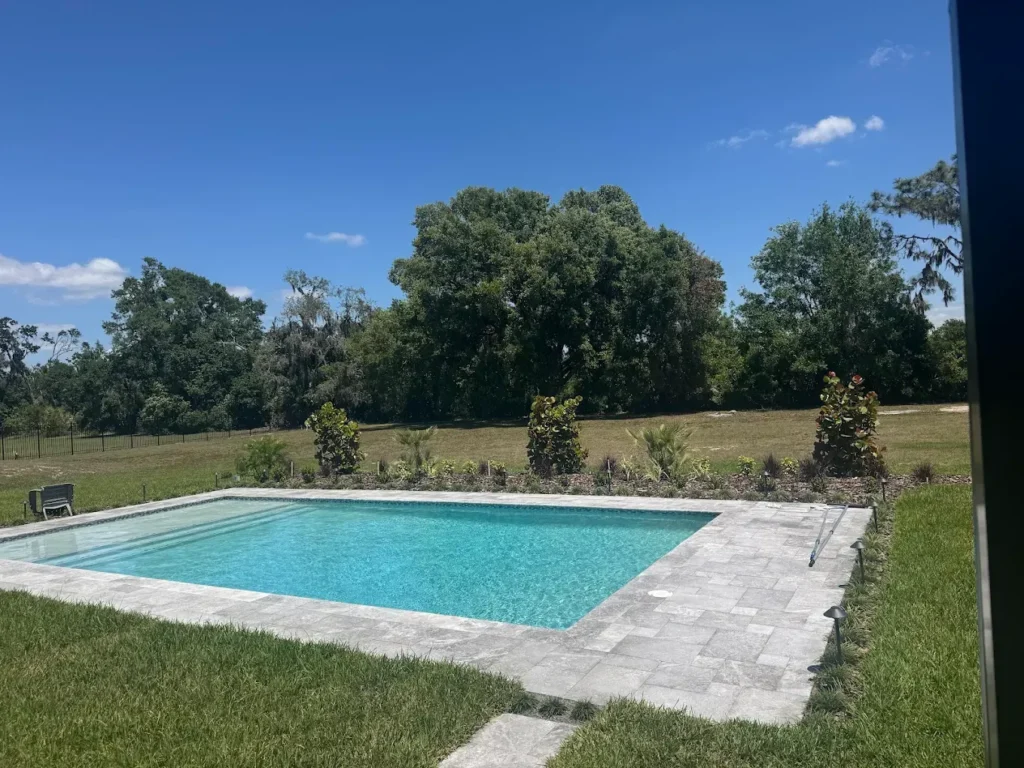 A rectangular backyard pool with travertine coping surrounded by open lawn and trees under a clear sky.