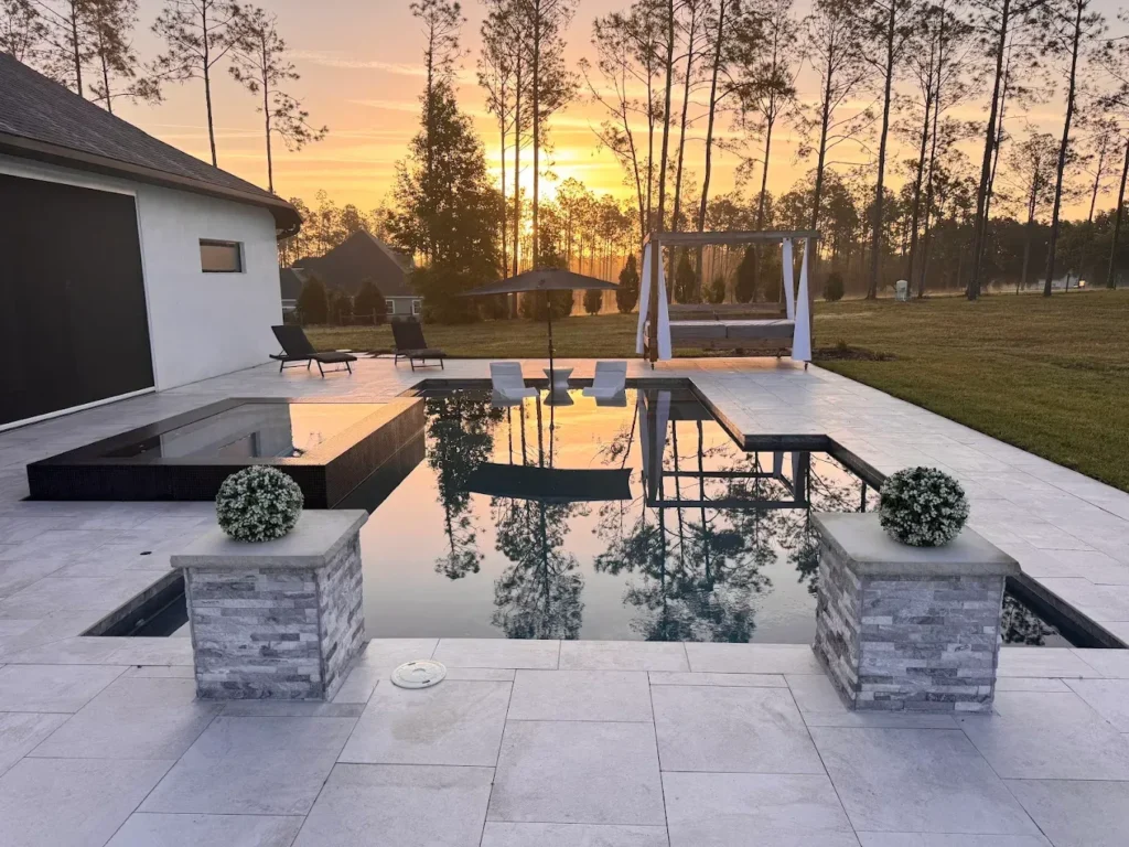 A modern geometric pool with a raised dark-tiled spa reflects a golden sunset behind a stone patio.