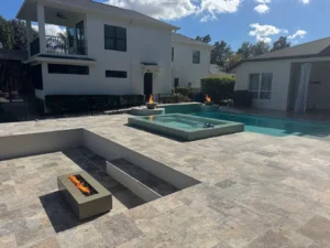 A raised rectangular spa sits beside a rectangular pool on a travertine patio with two fire bowls and a sunken fire pit area.