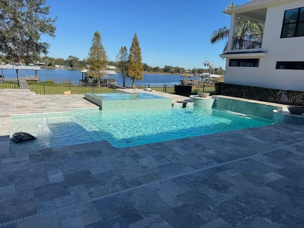 A modern geometric pool with a raised spillover spa sits on a travertine patio overlooking a lake.