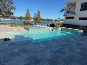 A modern geometric pool with a raised spillover spa sits on a travertine patio overlooking a lake.