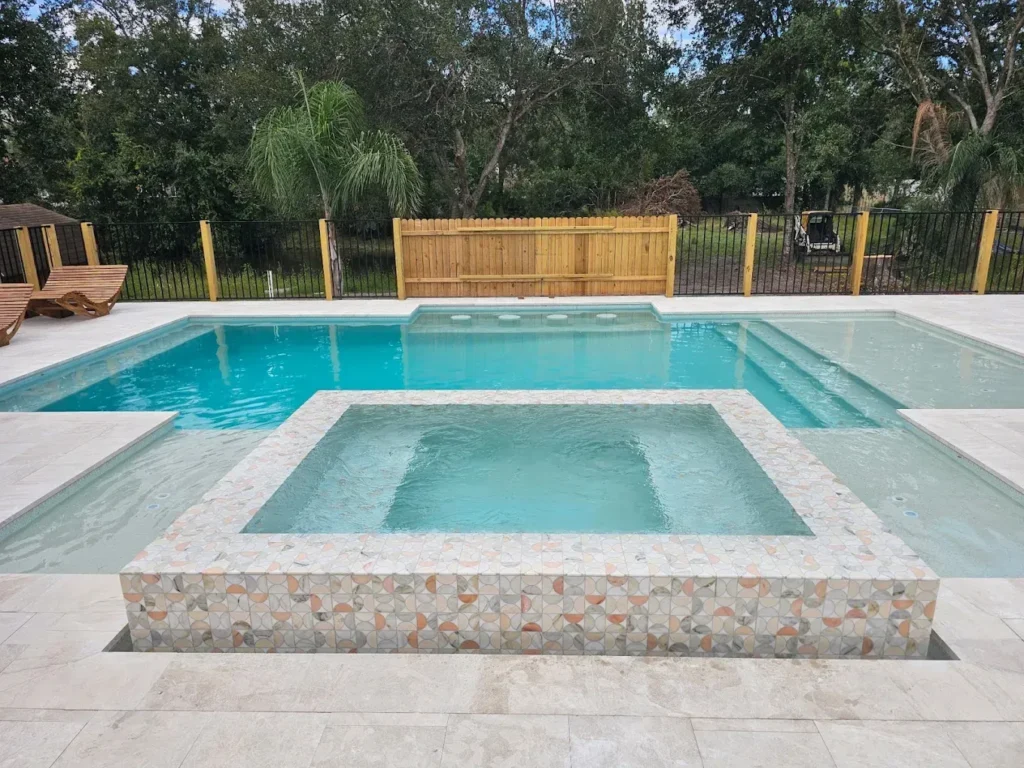 Rectangular pool with a raised mosaic-tiled square spa and tanning ledge on a travertine patio.