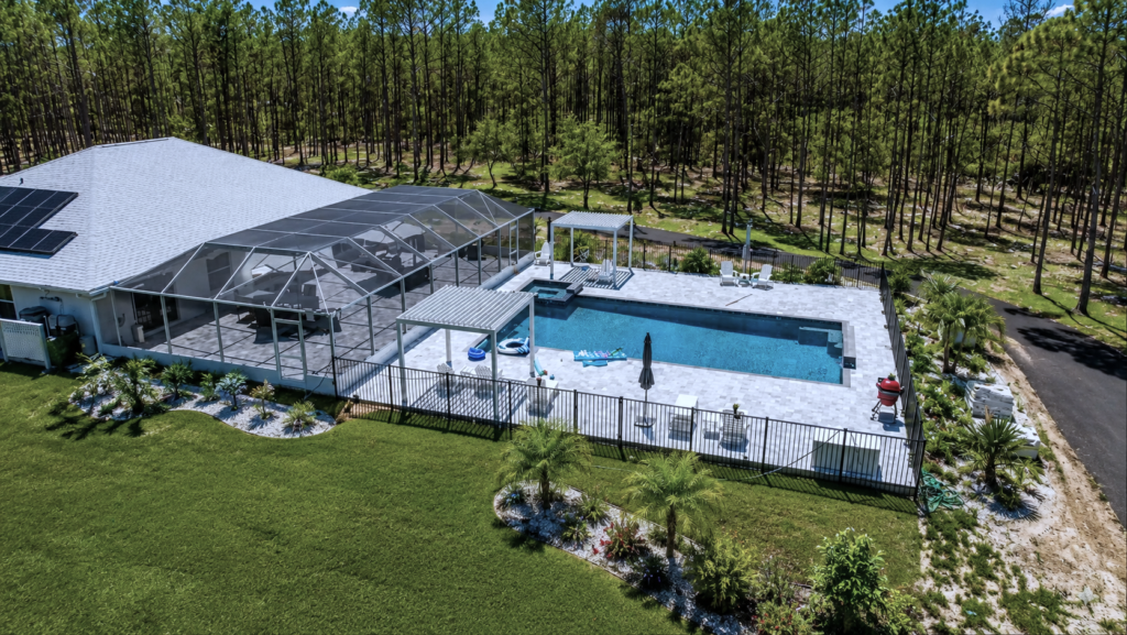Aerial view of a rectangular pool with a raised spa in a screened-free paver patio surrounded by pine woods.
