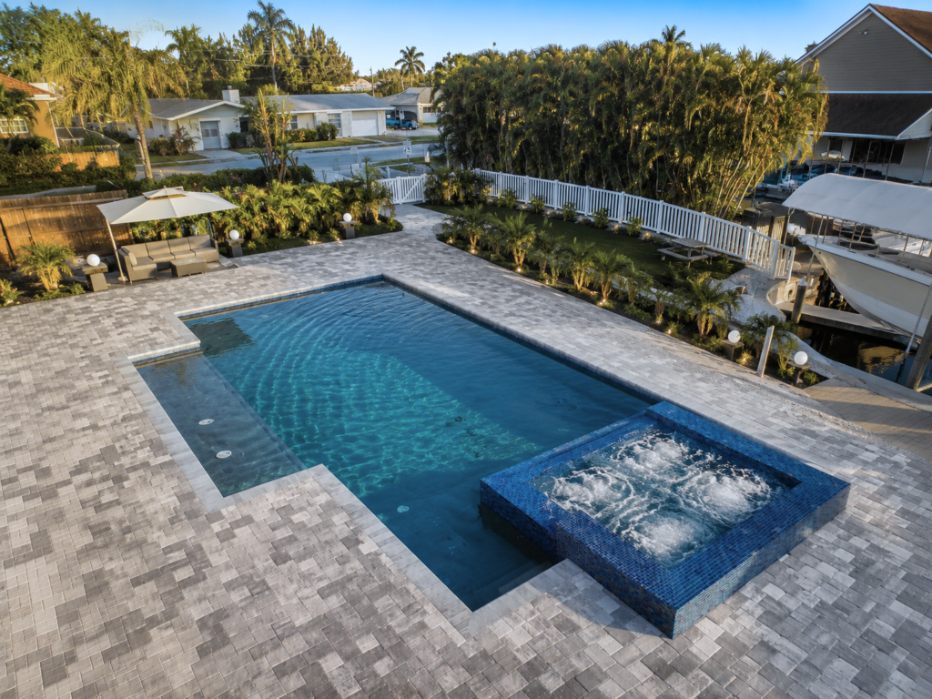 Aerial dusk view of a rectangular pool with a tiled raised spa and paver deck beside a canal.