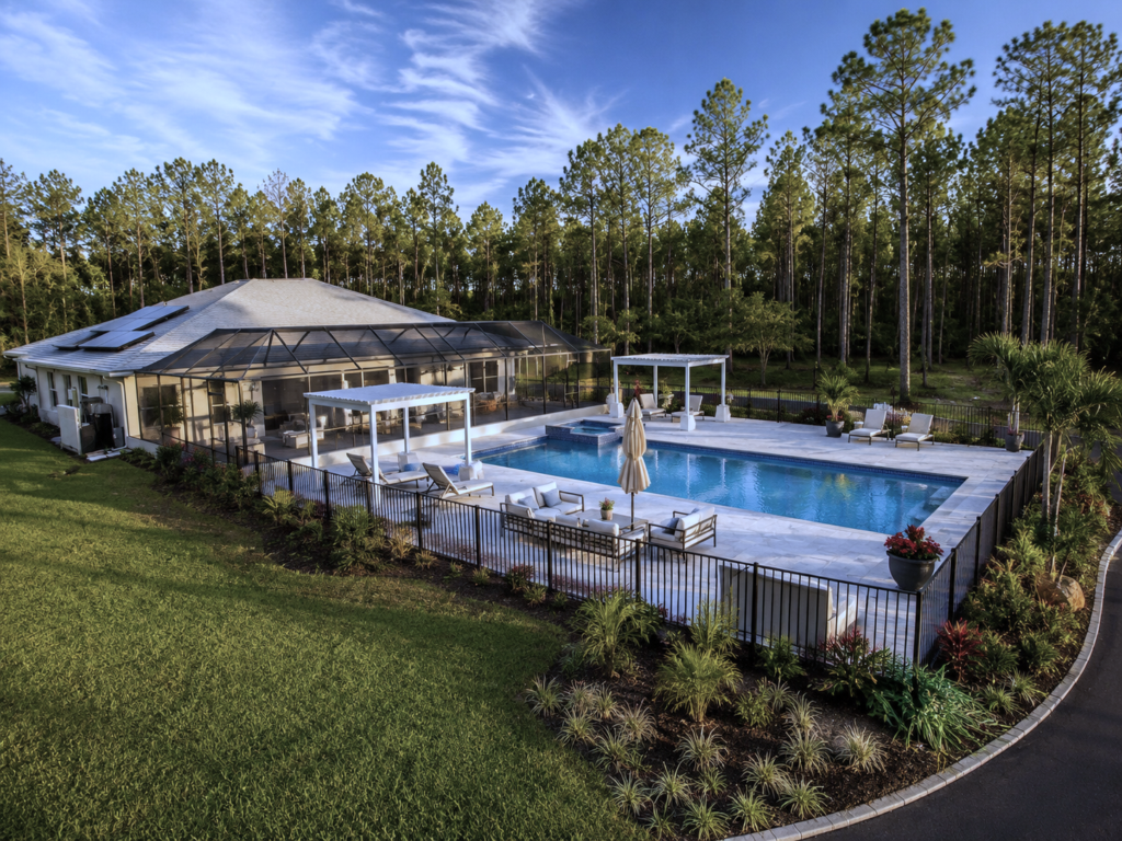 Aerial view of a rectangular backyard pool with attached raised spa, paver deck, and pergolas, surrounded by tall pines.
