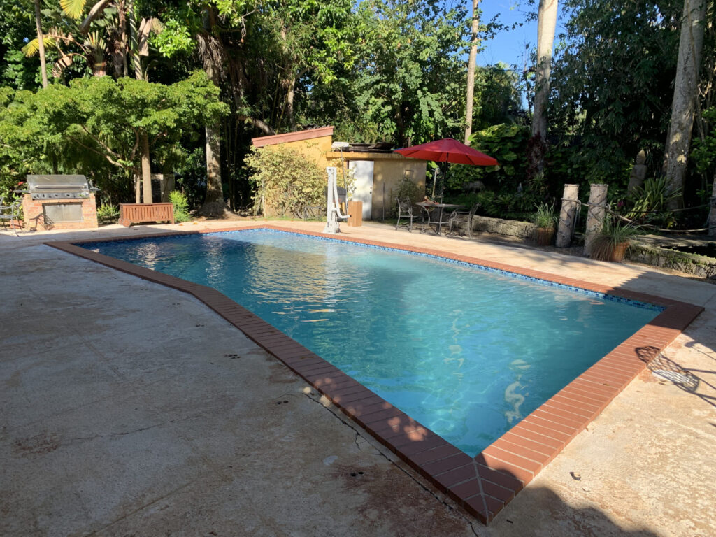 An L-shaped backyard pool with brick coping surrounded by mature trees and a patio with a grill and umbrella table.