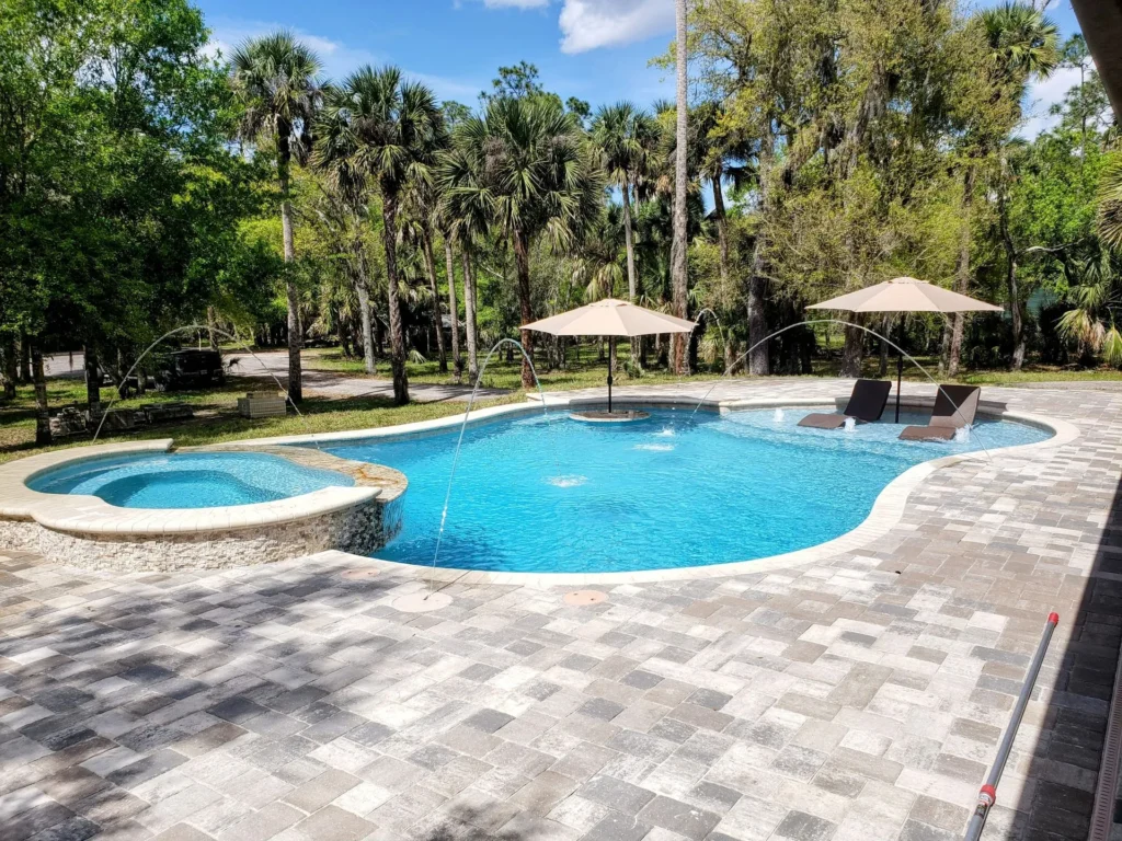 A freeform pool with a raised spa, deck jets, and in-pool loungers set on a paver patio surrounded by palm trees.
