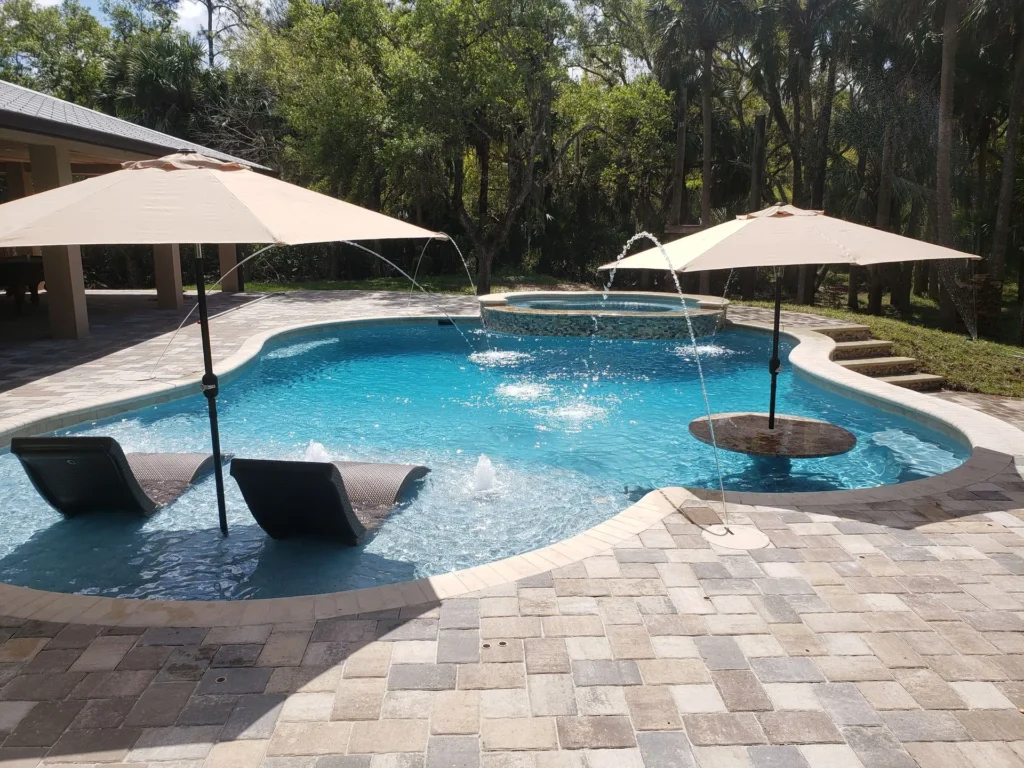 Freeform pool with in-water loungers, bubblers, and a raised spa surrounded by a paver patio and wooded backdrop.