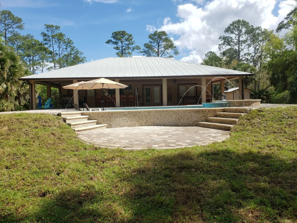 An elevated rectangular pool with stone-clad walls and deck jets sits on a raised patio behind a metal-roofed house surrounded by pine trees.