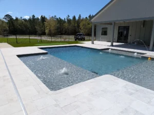 A rectangular pool with an attached sun shelf and bubbler jets sits on a travertine paver patio behind a modern farmhouse.