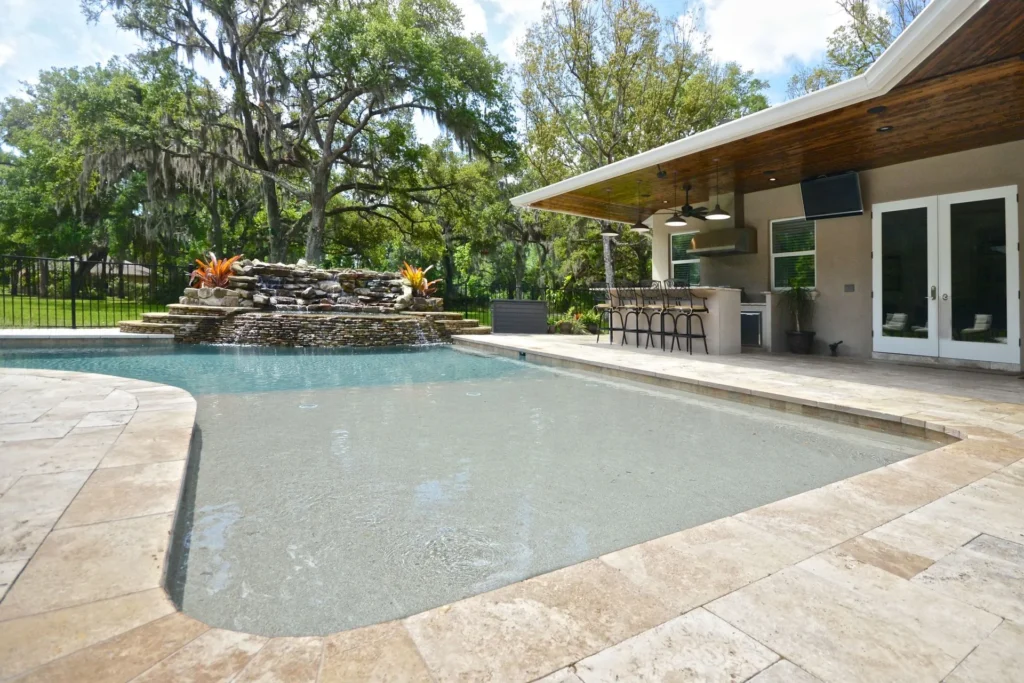 A freeform backyard pool with a rock waterfall feature beside a covered outdoor kitchen and lanai.