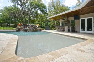 A freeform backyard pool with a rock waterfall feature beside a covered outdoor kitchen and lanai.