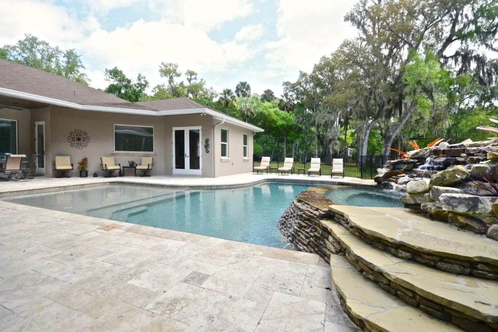 A freeform pool with a natural stone waterfall feature beside a travertine paver patio behind a single-story home.