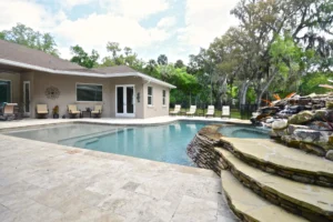 A freeform pool with a natural stone waterfall feature beside a travertine paver patio behind a single-story home.