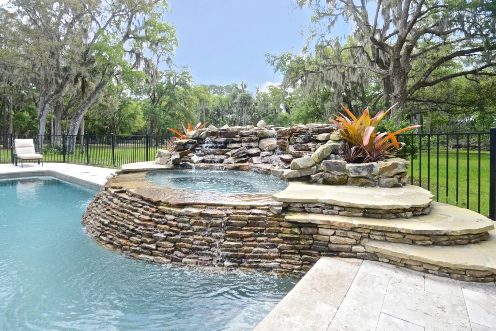A raised stone waterfall spa cascades into a freeform pool surrounded by trees.