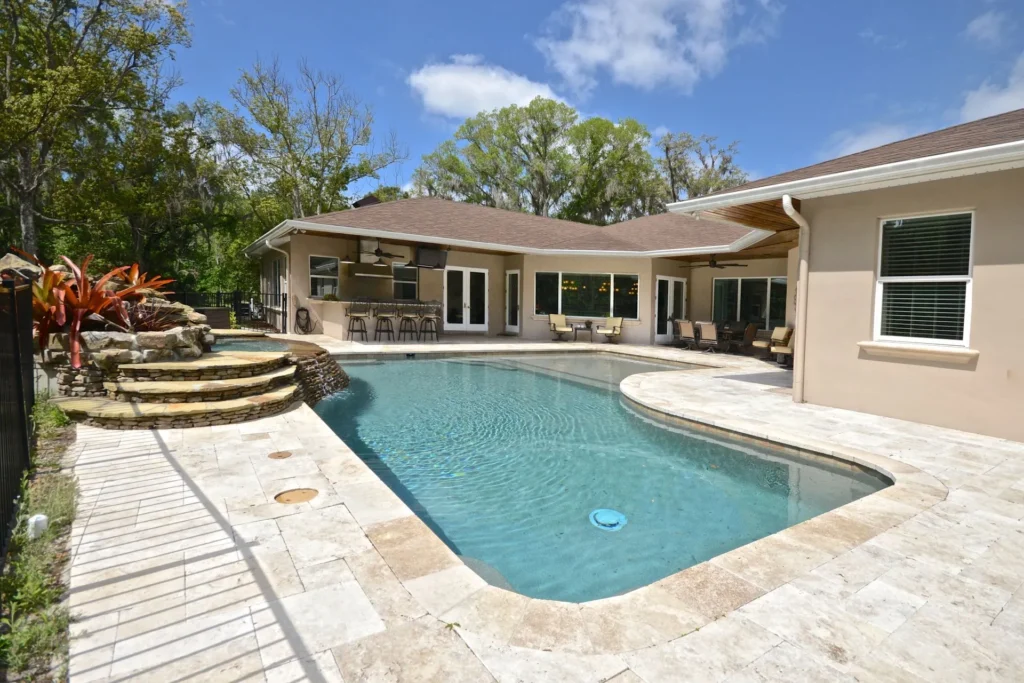 A freeform backyard pool with travertine decking, an outdoor kitchen, and a rock waterfall feature under a blue sky.
