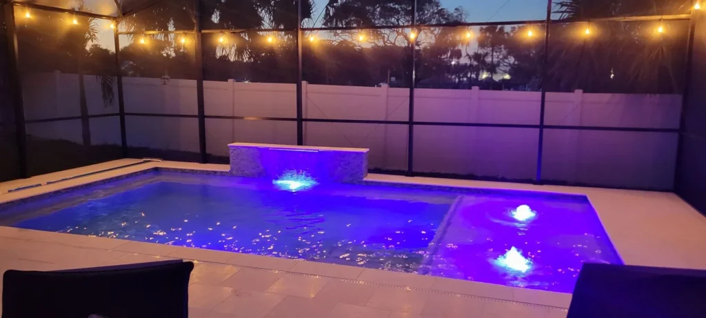 A small rectangular pool with vivid blue LED lighting and a raised stone water feature under a screened enclosure at night.