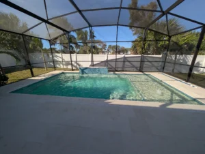 A rectangular pool with a raised wall water feature under a screened enclosure and travertine deck.