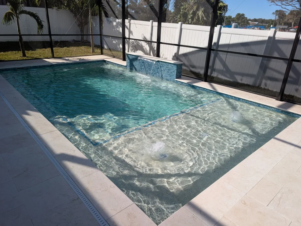 A rectangular pool with a shallow sun shelf and bubbler jets under a screened lanai.