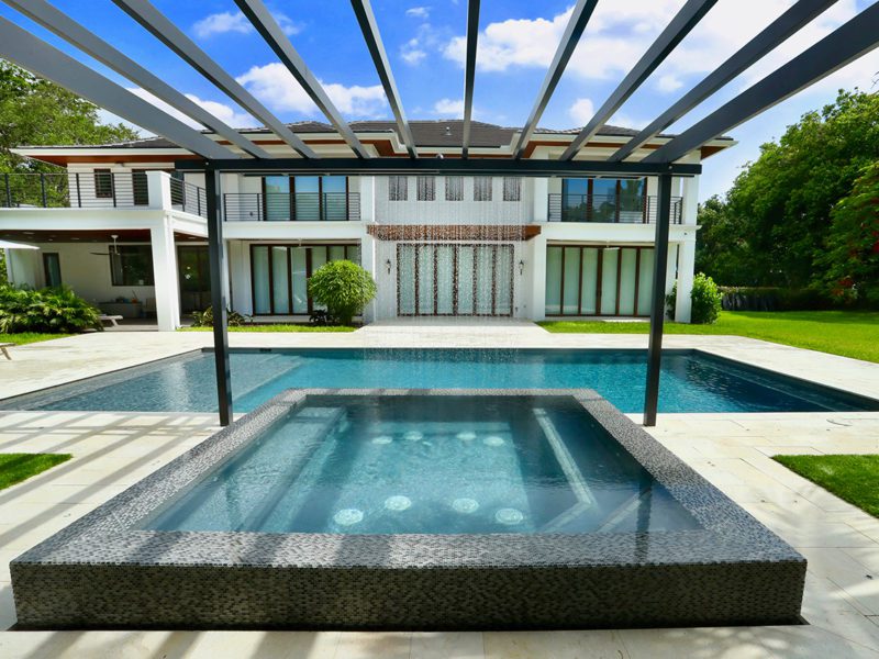 A raised mosaic-tiled spa with bubbler jets sits in front of a rectangular pool and a modern home with a water-wall feature, under a metal pergola.