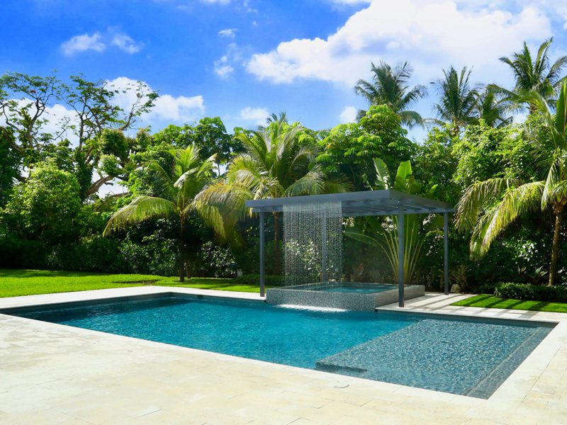 A rectangular pool with a tanning ledge and integrated spa beneath a modern rain-curtain water feature, surrounded by lush tropical landscaping.