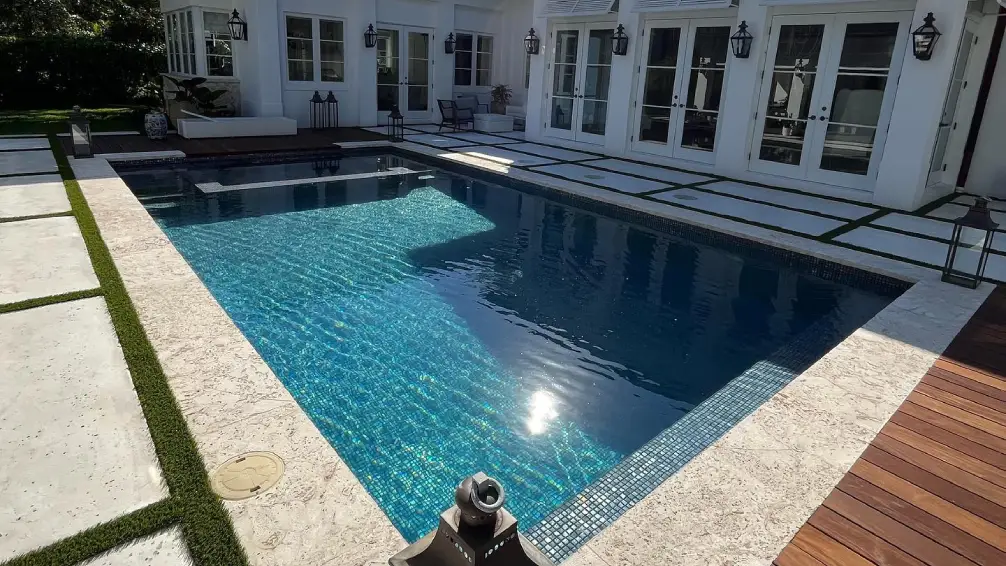 A rectangular pool with a dark tile waterline and travertine deck behind a white house.