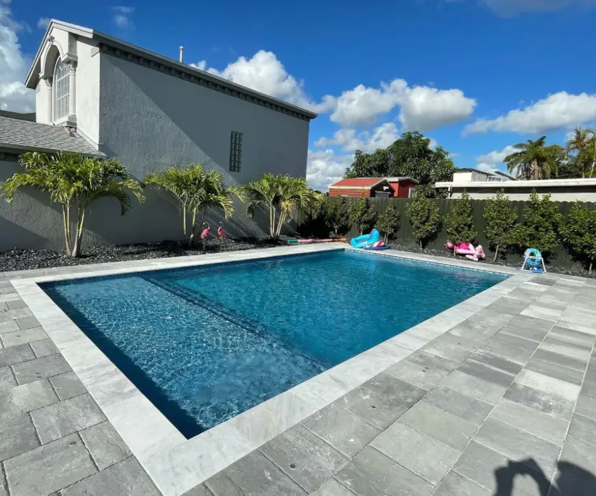 A rectangular backyard pool with travertine coping beside a stucco house and palm trees.