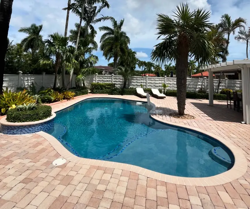 A freeform blue pool surrounded by brick pavers and palm trees in a fenced backyard.