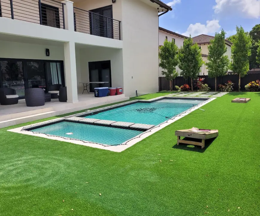 A rectangular backyard pool covered with a black safety net, surrounded by artificial turf and a two-story house.