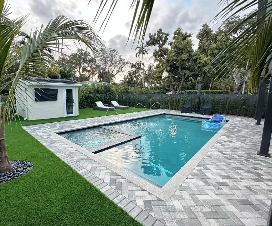 A rectangular backyard pool with a tanning ledge and water jets, surrounded by paver decking, turf, and palm trees at dusk.