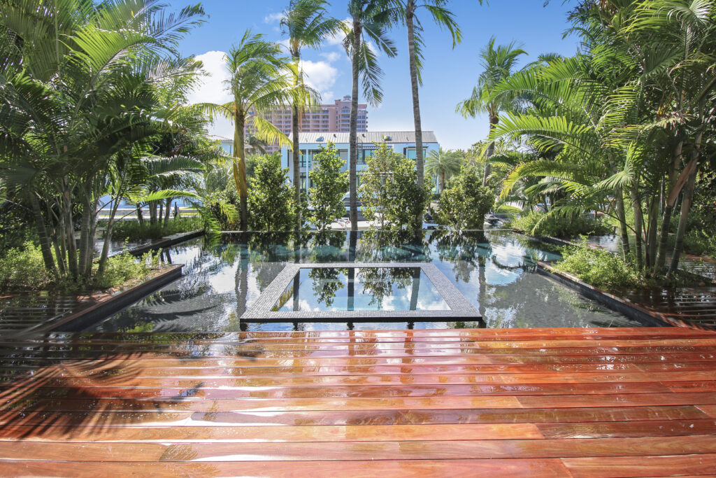 A long reflecting pool with a raised square tile island, framed by palm trees and a wet wooden deck with a resort building in the background.
