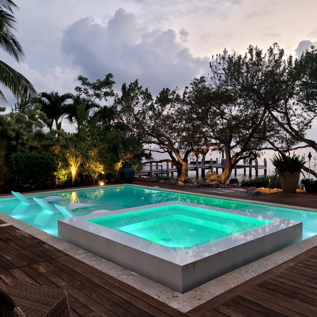 A rectangular pool with a raised glowing spa on a wood deck at dusk overlooking a canal.