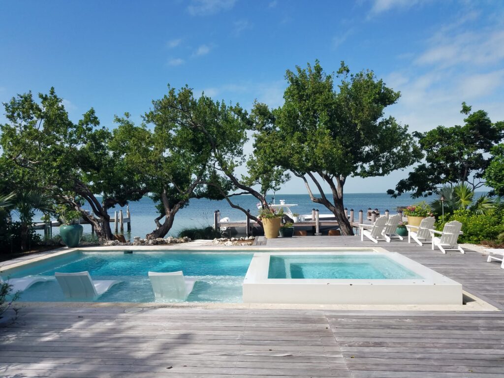A small rectangular pool with in-water loungers and a raised white spa sits on a wooden deck overlooking the ocean.
