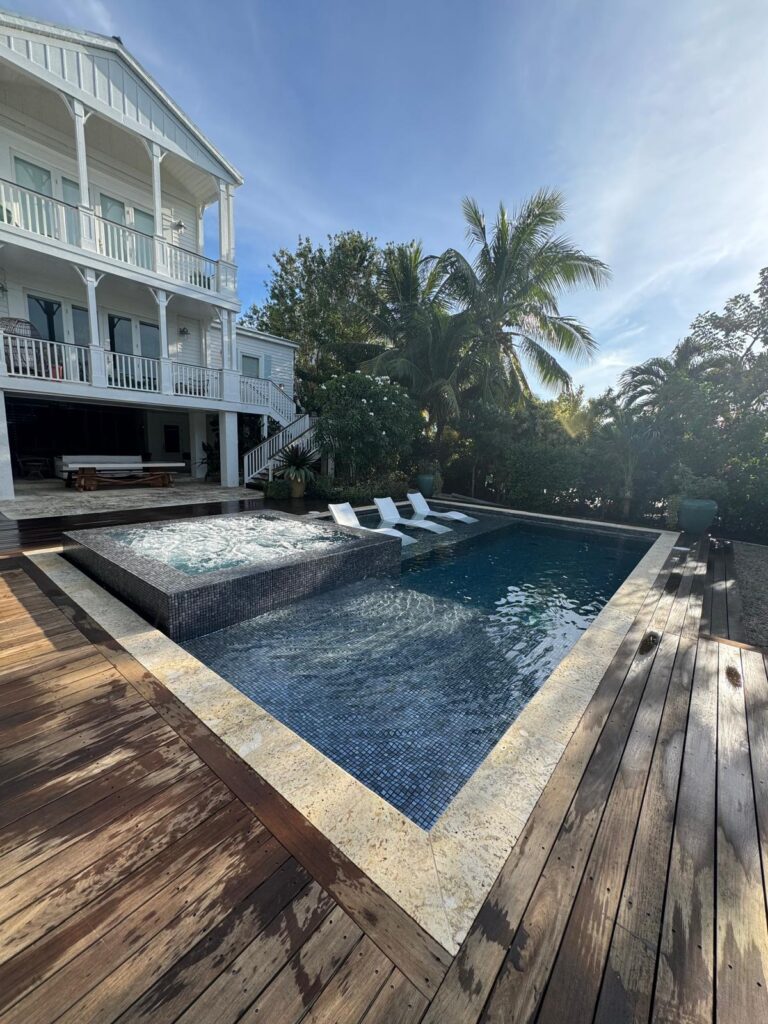 A tiled rectangular pool with a raised spa and built-in loungers on a wood deck beside a coastal-style home.