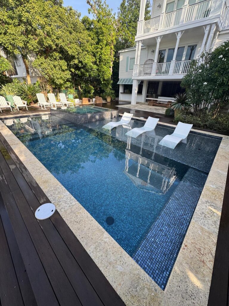A rectangular pool with blue mosaic tile interior, in-water lounge chairs, and a raised spa in front of a multi-story white house.
