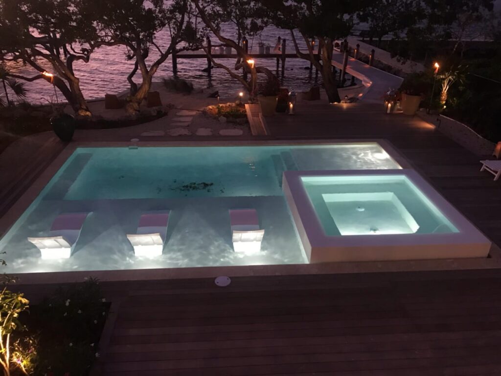 A rectangular pool with in-water loungers and a raised spa lit up at night beside a waterfront deck.