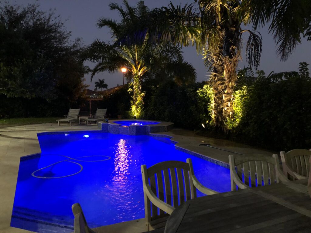 A freeform backyard pool with vivid blue LED lighting and an attached raised spa, surrounded by palm trees at night.