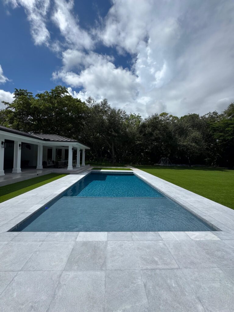 A long rectangular pool with light stone coping sits beside a covered patio under a partly cloudy sky.