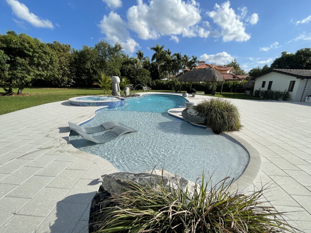 A freeform pool with a raised spa, in-pool loungers, and beach entry surrounded by a paver patio and tropical landscaping.