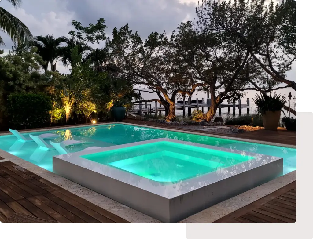 A modern rectangular pool with an elevated raised spa glowing turquoise at dusk beside a waterfront dock.