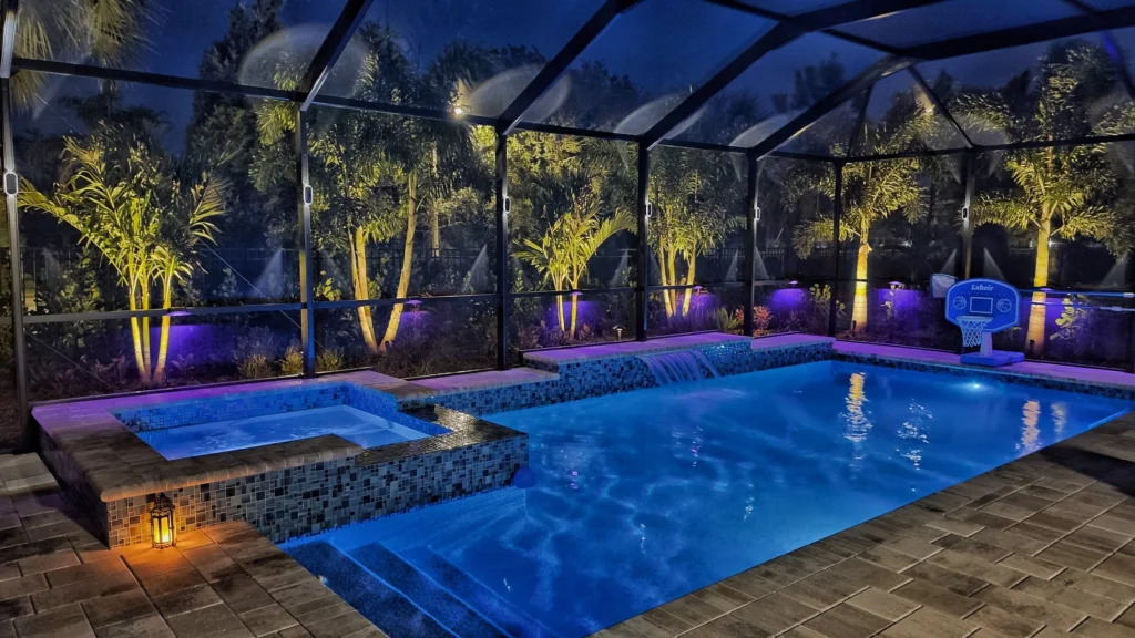 A screened Florida lanai at night with a rectangular pool lit blue, an attached raised spa spilling over a tiled wall, and a poolside basketball hoop.