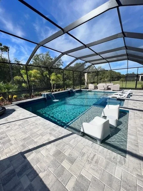 A screened backyard pool with a sun shelf, white in-water chairs, and water spouts under a birdcage enclosure.