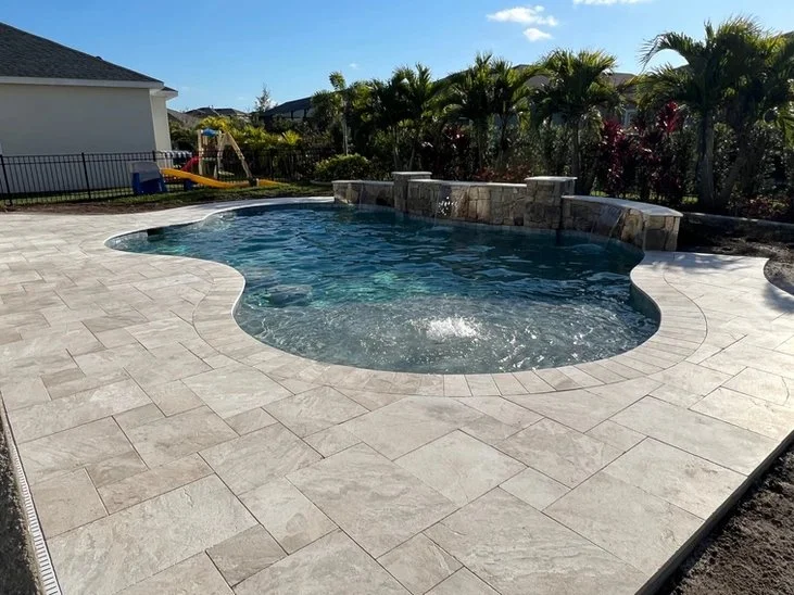 A freeform backyard pool with a bubbler fountain, stone accent wall, and travertine deck, with a play set visible in the yard.