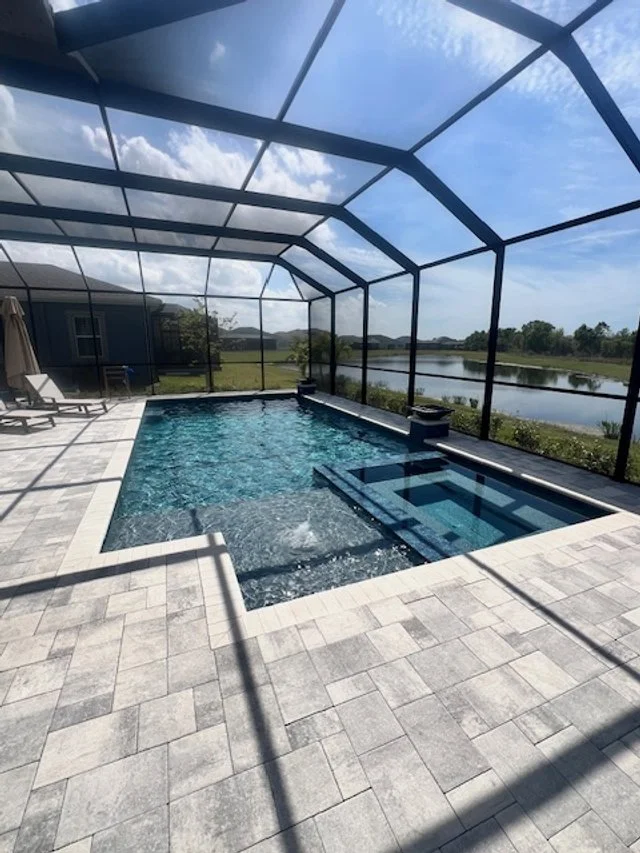 A screened lanai pool with an integrated spa and paver deck overlooking a pond.