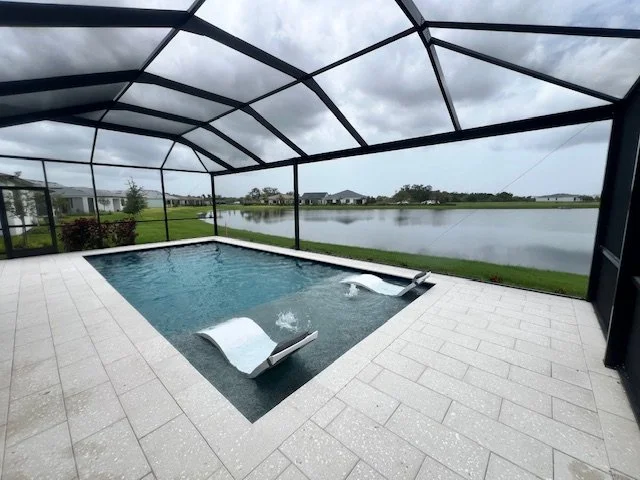 A screened rectangular pool with two in-water loungers overlooking a lake under a cloudy sky.