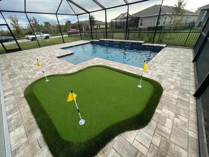 Small L-shaped pool with raised wall under a screened enclosure next to an artificial putting green.